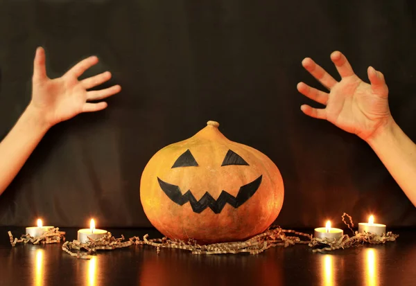 Mother`s and child`s hands next to Halloween pumpkin in disposable protective mask on the table with burning candles. Black background. Concept of Halloween party during coronavirus pandemic