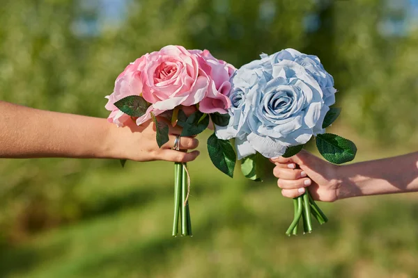 Two hands hold two bouquets of blue and pink roses. The image is surrounded by vegetation.