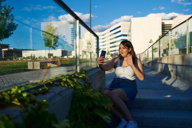 Video konferansında akıllı telefonuyla konuşan bir kadın. Park basamaklarında oturuyor. Güneşli bir gün..