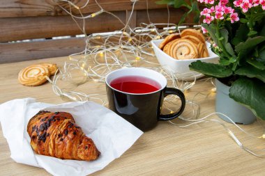 Taze kruvasan ve gül çaylı siyah bardak. Arka planda pembe çiçekler.