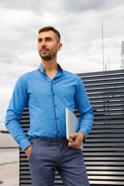 Mavi gömlekli genç bir adam dizüstü bilgisayar tutuyor..