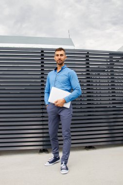 Mavi gömlekli genç bir adam dizüstü bilgisayar tutuyor..