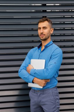 Mavi gömlekli genç bir adam dizüstü bilgisayar tutuyor..