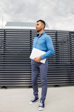 Mavi gömlekli genç bir adam dizüstü bilgisayar tutuyor..