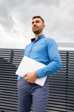 Mavi gömlekli genç bir adam dizüstü bilgisayar tutuyor..