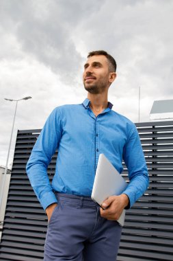 Mavi gömlekli genç bir adam dizüstü bilgisayar tutuyor..