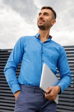 Mavi gömlekli genç bir adam dizüstü bilgisayar tutuyor..