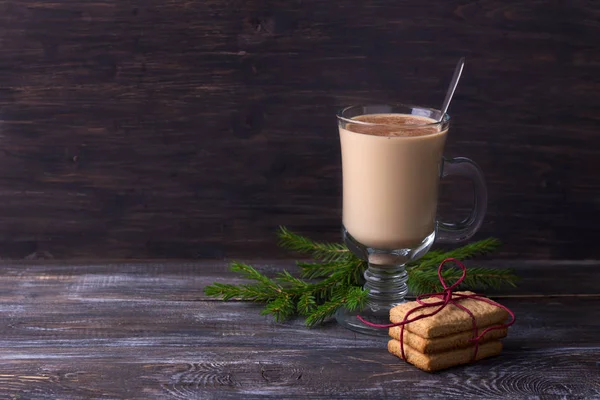Spicy warming tea with milk in a glass cup with cinnamon, gingerbread cookie tied with ribbon and Christmas tree branches on a wooden table