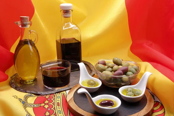 Products from Spain, Still life, board with bowl of mixed olives, 3 porcelain spoons with oil and olives, bottle and glass red wine and extra virgin olive oil on tray, on flag of spain, product photo