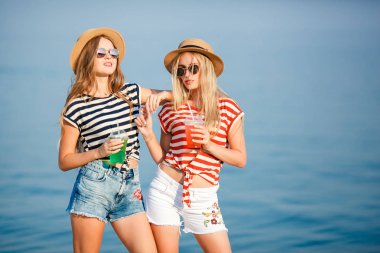 Trendy iki genç güzel kız portresi Denizi üzerinde bir kot gömlek gömlek, plaj şapka, güneş gözlüğü şerit. iki arkadaş kokteyller elleri ile denizde eğleniyor.
