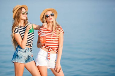 Trendy iki genç güzel kız portresi Denizi üzerinde bir kot gömlek gömlek, plaj şapka, güneş gözlüğü şerit. iki arkadaş kokteyller elleri ile denizde eğleniyor.
