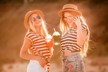 Trendy iki genç güzel kız portresi Denizi üzerinde bir kot gömlek gömlek, plaj şapka, güneş gözlüğü şerit. iki arkadaş kokteyller elleri ile denizde eğleniyor. Gençlik ve mutluluk kavramı.