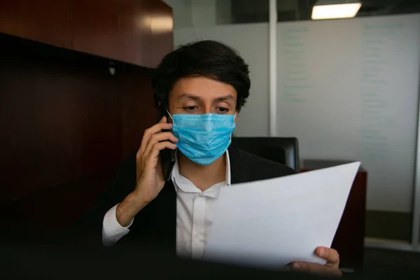 Telefonda konuşan bir adamın stok fotoğrafı. Virüsü önlemek için koruyucu bir maske takıyor. Ofiste çalışıyor.