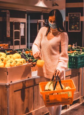 Güzel esmer kız süpermarketteki alışveriş sepetine tatlı biber ekliyor. Açık renk kazak, beyaz kot pantolon ve mavi tıbbi maske giyiyor.