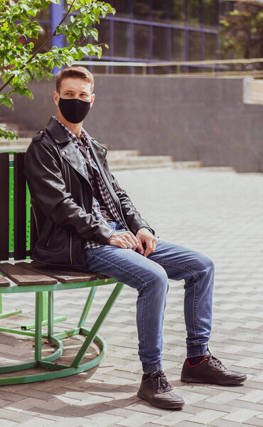 Caucasian men sits on the bench and turned his head to the left. He is looking far away. Sits near the shop in the city. Dressed into jeans, T-shirt, leather jacket and black protective mask.