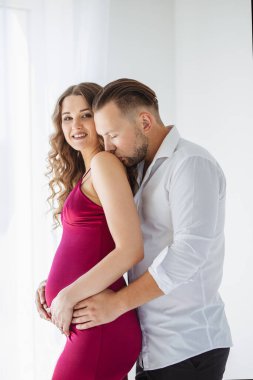 Gece elbisesi giymiş güzel kıvırcık, hamile bir kız kocasıyla kucaklaşıyor. Koyu pembe bir gece elbisesi var ve adam güzel beyaz bir gömlek giyiyor ve omzunu nazikçe öpüyor.