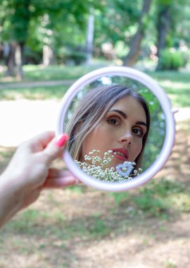 Bir kızın yüzü pembe bir manikürle elinde tuttuğu küçük pembe bir aynaya yansıyor. Bir parkta ağacın yanında duruyor. (Sadece yüze odaklan.))