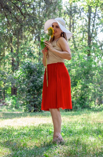 Beyaz şapkalı güzel bir kadın elinde sarı bir ayçiçeğiyle parkta duruyor. Hava güneşli ve dışarıda yaz var.