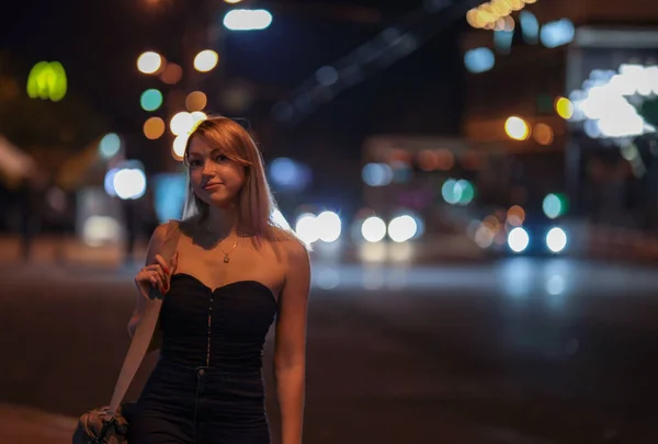 girl stands on the side of the road at night in a black corset and with a bag on her shoulders. City and cars on blurred background. Picture has dark night  tone.