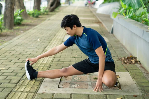 Asyalı genç fitness koşucusu parkta koşmadan önce bacağını çırpıyor.