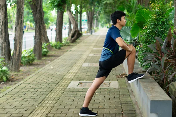 Asyalı genç fitness koşucusu parkta koşmadan önce bacağını çırpıyor.