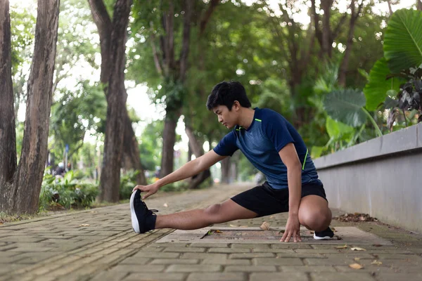 Asyalı genç fitness koşucusu parkta koşmadan önce bacağını çırpıyor.