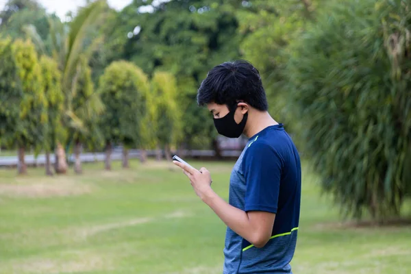 Młody Fitness Człowiek Maską Parku Słuchawkach Bezprzewodowych Smartwatch Sprawdzanie Telefonu — Zdjęcie stockowe