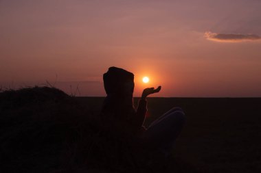 Kapüşonlu, elleri güneşe yükselmiş bir kızın karanlık silueti.