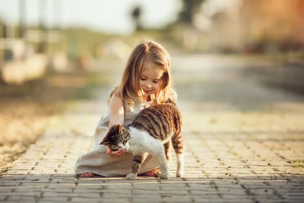 Маленькая босоногая девочка в сером льняном платье гладит кошку. Солнечный летний вечер в деревне. Деревенский фон. Изображение с избирательным фокусом, тонизирующим и шумовым эффектом . — стоковое фото