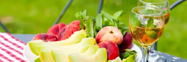 Summer party outdoor. Romantic picnic for a couple. Two glasses of cool wine served with mint and lime. Fresh melon and peach as simple light meal. Tasty low calories dessert. Green background. Banner