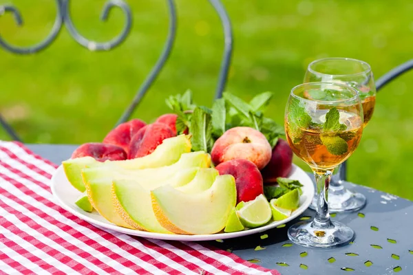 Summer party outdoor. Romantic picnic for a couple. Two glasses of cool wine served with mint and lime. Fresh melon and peach as simple light meal. Tasty low calories dessert. Green background.