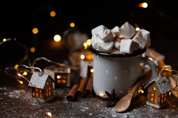 Winter hot drink: hot chocolate with marshmallows, cinnamon and anise. Christmas lights in the shape of houses as decor. Decorative snow, dark wooden background. Festive mood, cozy home atmosphere