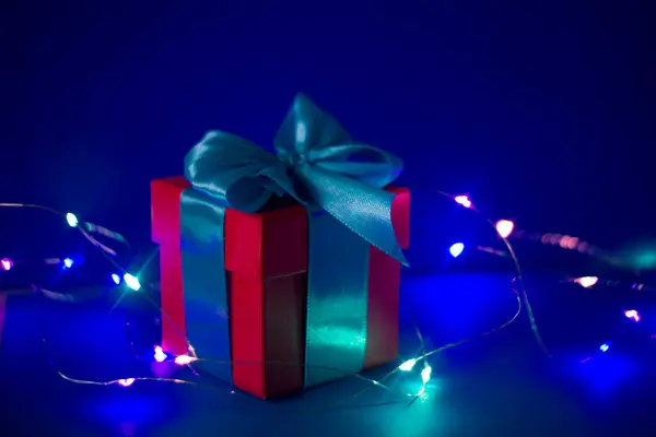Mavi arka planda mavi kurdeleli kırmızı hediye kutusu ve renkli ışıklar. — Stok fotoğraf