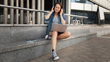 Genç ve güzel bir kız laptopun başına oturur ve korkulukta oturup müzik dinler.