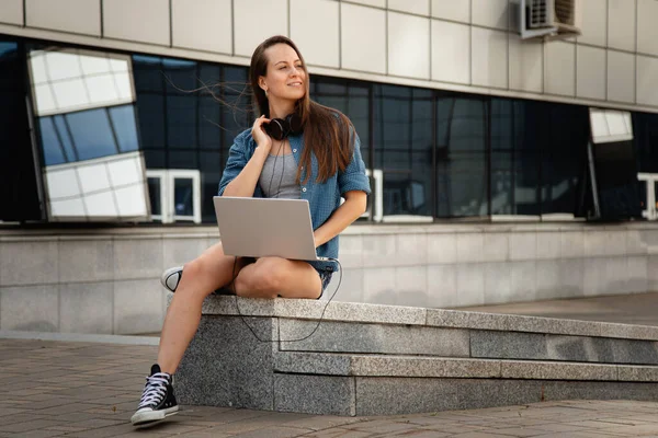 young beautiful girl sit at work at the laptop and listen to music while sitting on the parapet