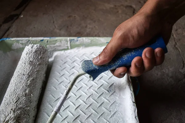 Hand of a house painter with a paint roller. Paint roller with white paint in hand, lowers into a tray with paint, on a concrete floor background