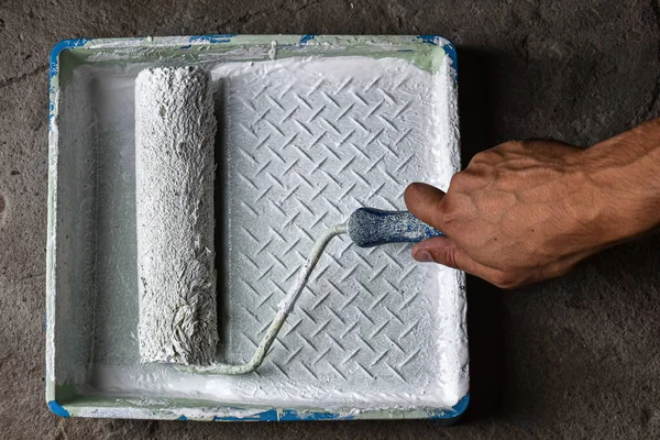 Hand of a house painter with a paint roller. Paint roller with white paint in hand, lowers into a tray with paint, on a concrete floor background