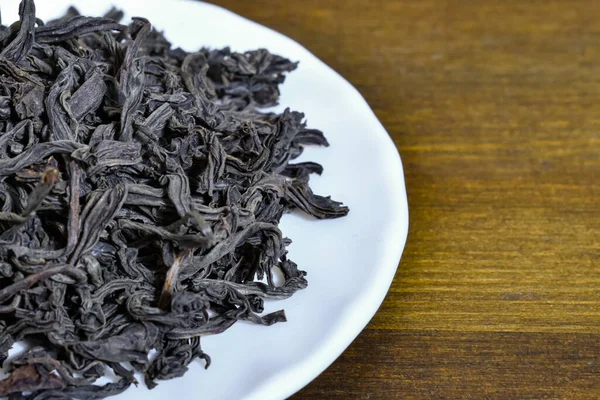 Large-leaf black tea on a white plate on a brown wooden background. Close-up Dried large black tea leaves in a white saucer on a wooden surface. Selective focus.