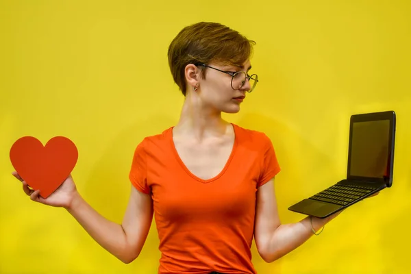 A young woman with glasses makes a choice, holding a red heart in one hand and a laptop in the other. Isolated on a yellow background. Concept: love, technology, long distance relationships, learning