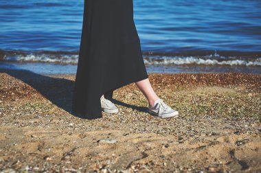Kum üzerinde beyaz spor ayakkabı kadın ayaklarda. Sahil boyunca yürüyerek. Siyah elbise giyen kadın. Retro tonlu görüntü
