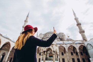 Genç Avrupalı kadın İstanbul, Türkiye 'de selfie portresi çekiyor. Kız kış İstanbul 'unda yürüyor. Sarışın, sonbahar günü bir caminin arka planında telefonla fotoğraf çekiyor..