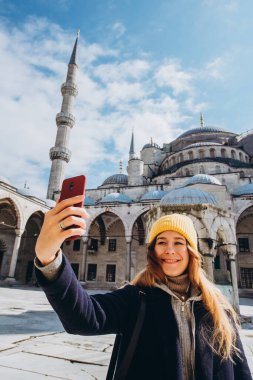 Genç Avrupalı kadın İstanbul, Türkiye 'de selfie portresi çekiyor. Kız kış İstanbul 'unda yürüyor. Sarışın, sonbahar günü bir caminin arka planında telefonla fotoğraf çekiyor..