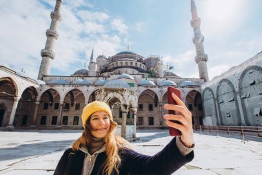 Genç Avrupalı kadın İstanbul, Türkiye 'de selfie portresi çekiyor. Kız kış İstanbul 'unda yürüyor. Sarışın, sonbahar günü bir caminin arka planında telefonla fotoğraf çekiyor..