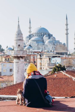 Sarışın bir kadın çatıda cami manzaralı bir kediyi okşuyor, İstanbul, Türkiye. İstanbul 'da güneşli bir sonbahar gününde şapkalı bir kız çatıda oturuyor. Gezgin kız kışın İstanbul 'da yürüyor.