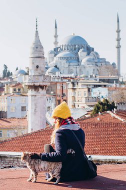 Sarışın bir kadın çatıda cami manzaralı bir kediyi okşuyor, İstanbul, Türkiye. İstanbul 'da güneşli bir sonbahar gününde şapkalı bir kız çatıda oturuyor. Gezgin kız kışın İstanbul 'da yürüyor.