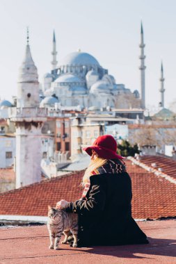Sarışın bir kadın çatıda cami manzaralı bir kediyi okşuyor, İstanbul, Türkiye. İstanbul 'da güneşli bir sonbahar gününde şapkalı bir kız çatıda oturuyor. Gezgin kız kışın İstanbul 'da yürüyor.