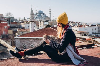 Sarışın bir kadın çatıda cami manzaralı bir kediyi okşuyor, İstanbul, Türkiye. İstanbul 'da güneşli bir sonbahar gününde şapkalı bir kız çatıda oturuyor. Gezgin kız kışın İstanbul 'da yürüyor.