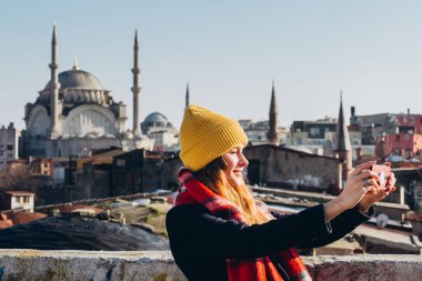 Sarışın kadın, İstanbul, Türkiye 'deki Grand Bazaar' ın çatısında telefonla fotoğraf çekiyor. Sarı şapkalı kız güneşli bir sonbahar gününde selfie çekiyor. Gezgin kız kışın İstanbul 'da yürüyor.