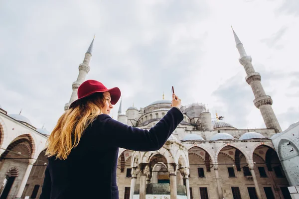 Genç Avrupalı kadın İstanbul, Türkiye 'de selfie portresi çekiyor. Kız kış İstanbul 'unda yürüyor. Sarışın, sonbahar günü bir caminin arka planında telefonla fotoğraf çekiyor..