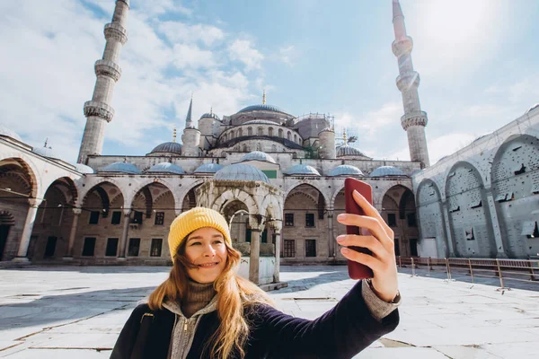 Genç Avrupalı kadın İstanbul, Türkiye 'de selfie portresi çekiyor. Kız kış İstanbul 'unda yürüyor. Sarışın, sonbahar günü bir caminin arka planında telefonla fotoğraf çekiyor..